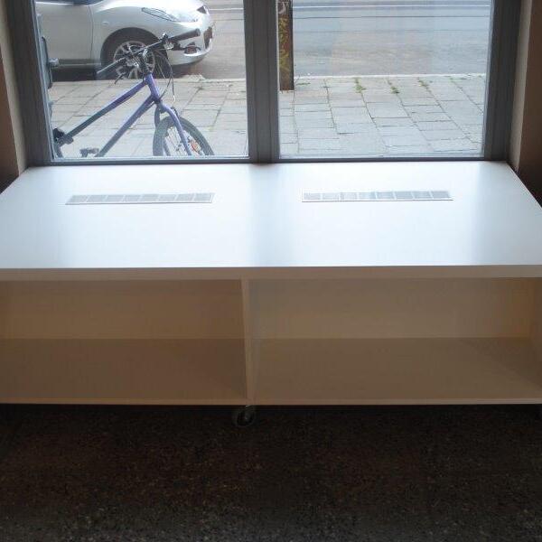 Ein weißes, maßgefertigtes Ladenregal auf Rollen, platziert vor einem Fenster, durch das ein Fahrrad und ein Auto sichtbar sind.