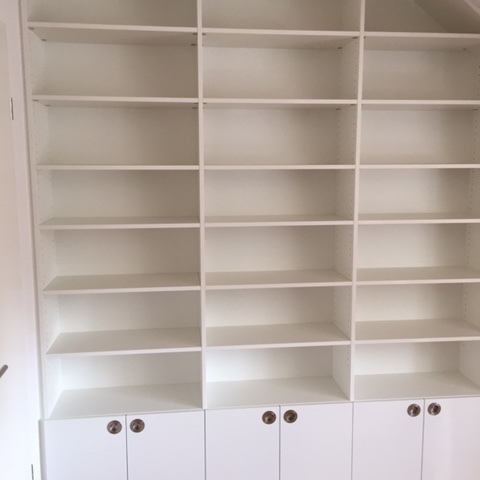 Ein leeres, weißes Regal nach Maß eingepasst in eine Dachschräge mit offenen Fächern und geschlossenen Schränken unten.