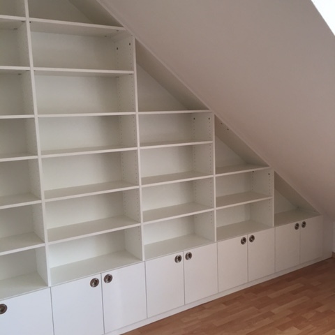 Weiße, maßgefertigte Regalwand, abgestuft und angepasst an eine Dachschräge, mit unteren Schranktüren.