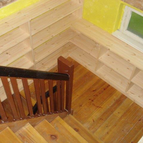 Maßangefertigtes Eckregal aus hellem Holz, montiert über einer Holztreppe neben einem Fenster.