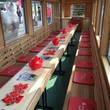 Innenansicht eines nach Maß ausgebauten Waggons, eingerichtet mit Tischen, roten Hockern und Werbematerialien.