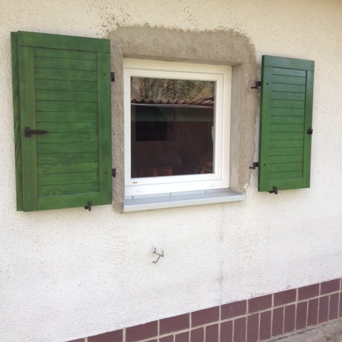 Grün gestrichene Fensterladen nach Maß, offen an einem Fenster mit weißem Rahmen und rustikaler Hauswand.