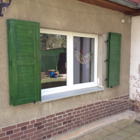 Ein großes Fenster mit offenen, grünen Fensterladen nach Maß, montiert an einer verputzten Hauswand.