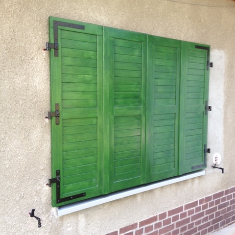 Geschlossene, leuchtend grüne Fensterladen nach Maß an einem Fenster, eingefasst von einer hellen Wand und einer Ziegelsockelleiste.