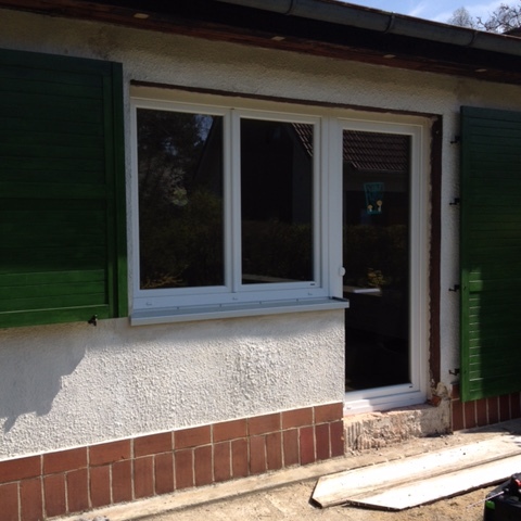 Grüne, offene Fensterladen nach Maß an einem weißen Fenster eines Hauses, unter einem blauen Himmel.