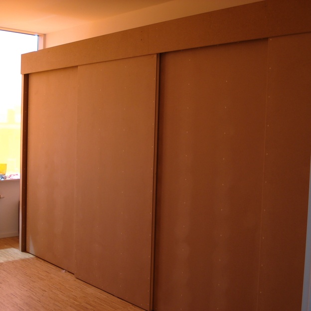 Maßgefertigtes Einbaumöbel mit großen Schiebetüren aus Holz in einem hellen Raum mit Parkettboden und bodentiefem Fenster.