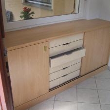 Helles Einbaumöbel mit Schiebetüren und offenen Schubladen, gefertigt aus Holz, in einem Raum mit Fliesenboden und bodentiefem Fenster.