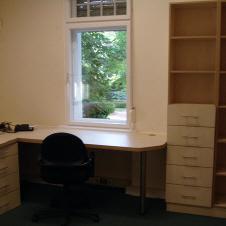 Helles Einbaumöbel mit großem Eckschreibtisch, mehreren Schubladen und einem offenen Regal in einem Büro mit Teppichboden und Fenster.