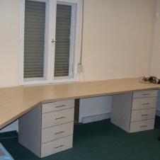 Eckschreibtisch aus hellem Holz mit zwei Schubladenelementen in einem Büro mit grünem Teppichboden und Fenster.