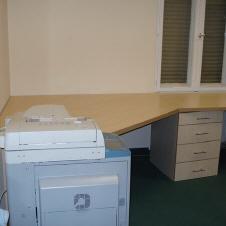 Maßgefertigter Eckschreibtisch aus hellem Holz mit Schubladenelementen in einem Büro mit grünem Teppichboden und Drucker im Vordergrund.