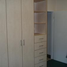 Maßgefertigter Einbauschrank aus hellem Holz mit mehreren Türen, Schubladen und offenen Regalfächern in einem Raum mit grünem Teppichboden.