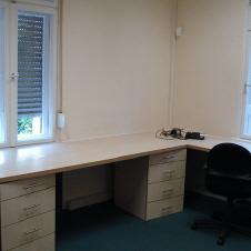 Maßgefertigter Eckschreibtisch aus hellem Holz mit mehreren Schubladenelementen in einem Büro mit grünem Teppichboden und zwei Fenstern.