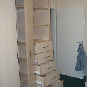 Maßgefertigter Einbauschrank aus hellem Holz mit offenen Schubladen und mehreren Regalfächern in einem Raum mit grünem Teppichboden.