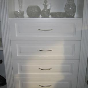 Maßgefertigter weißer Schrank mit vier Schubladen, verzierten Fronten und eleganten Metallgriffen, eingebaut in eine Wohnwand mit Glasdekor.