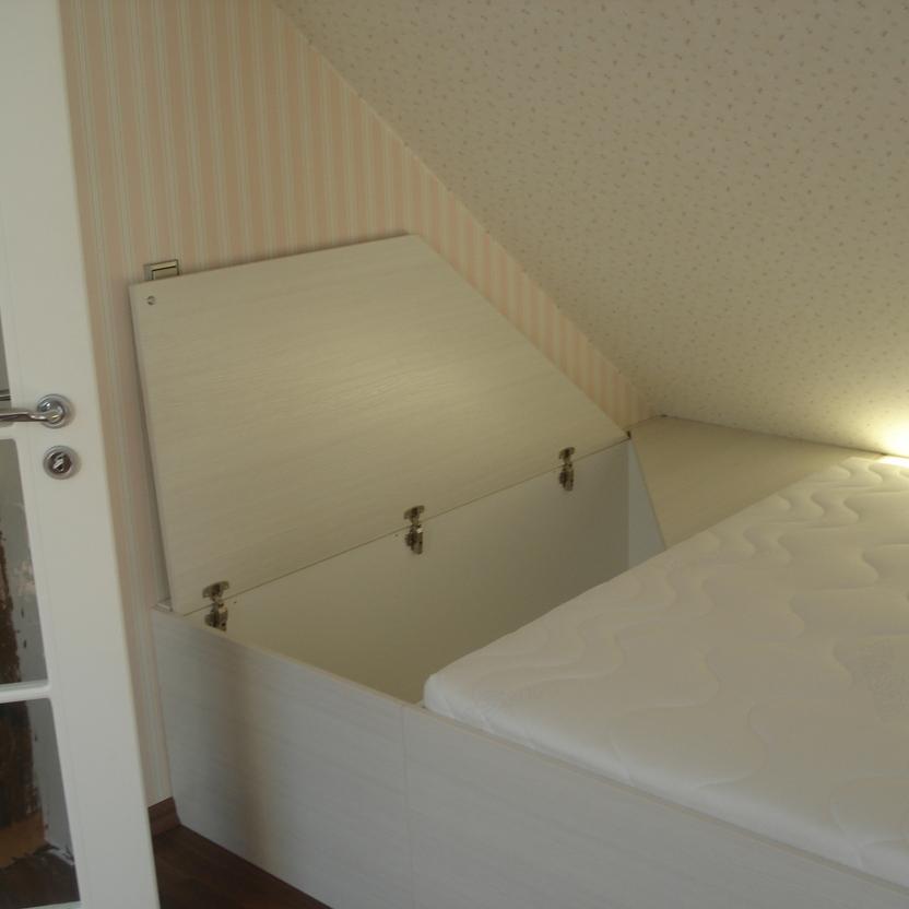 Maßgefertigtes Bett mit integriertem Stauraum und Klappe zum Öffnen, perfekt angepasst an eine Dachschräge in einem gemütlich eingerichteten Raum.